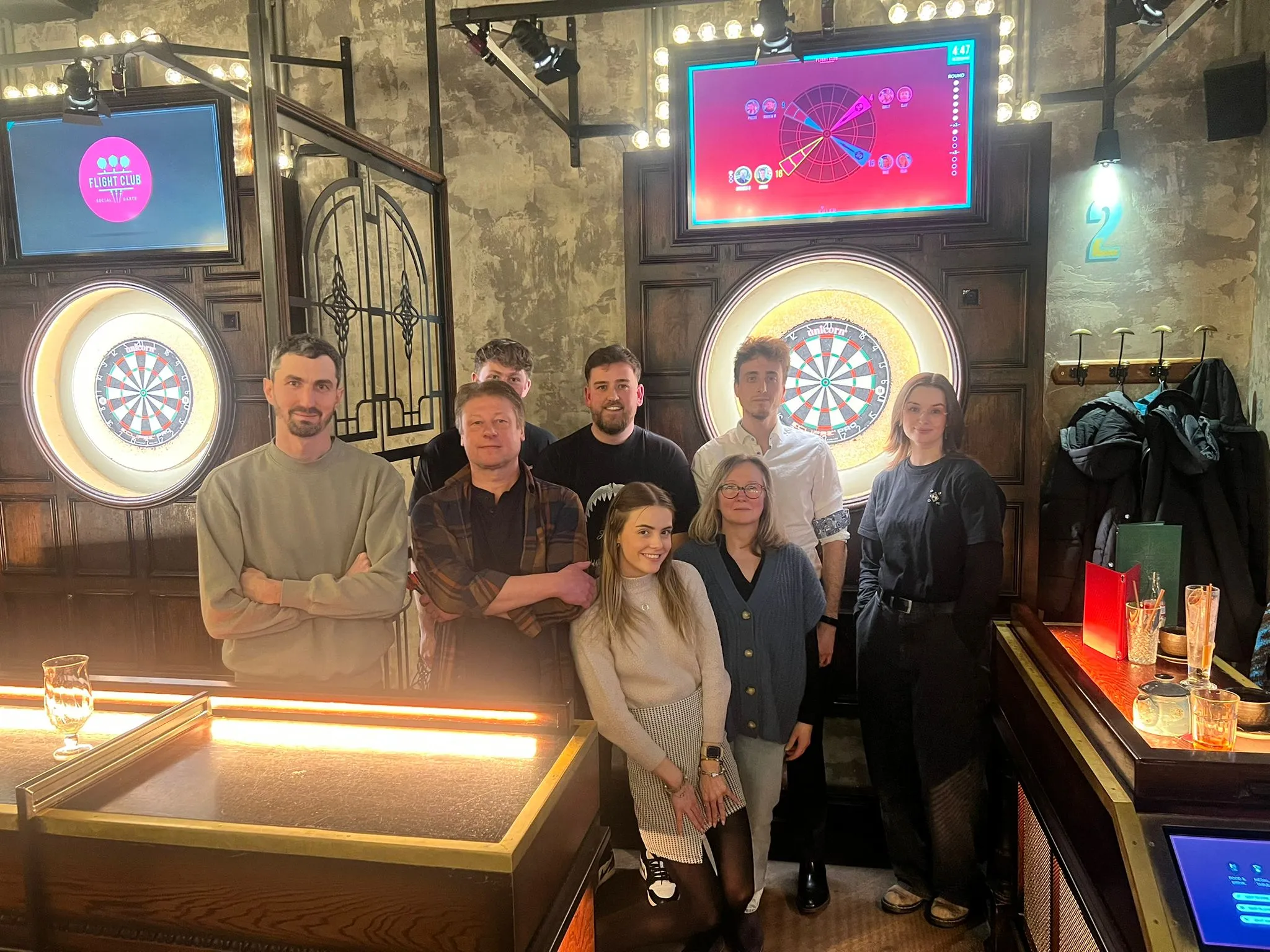 The eba team standing together in front of the illuminated dart boards at Flight Club Manchester.