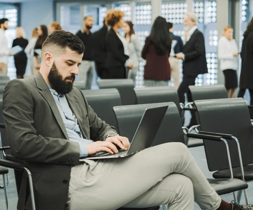 A director working on a laptop in an airport departure lounge while other travellers wait behind him.