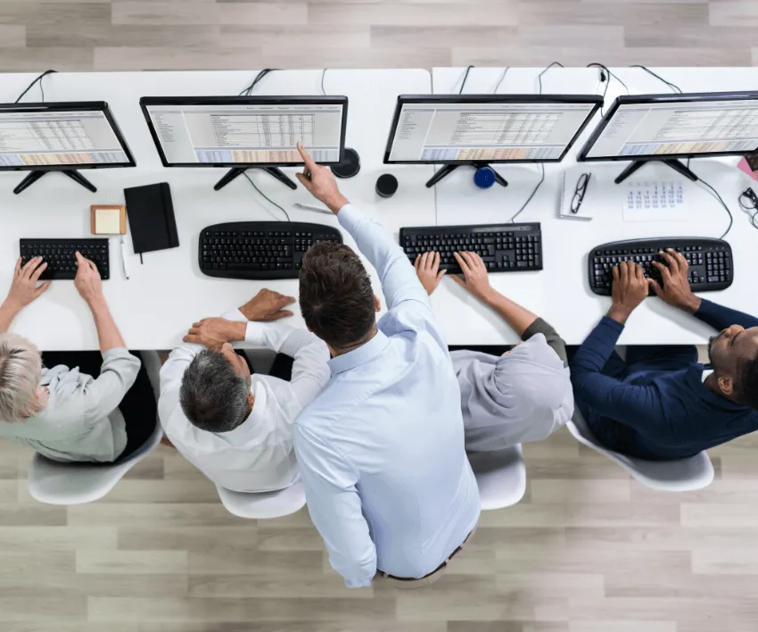 An overhead view of a team working at a row of desktop computers along one long desk.