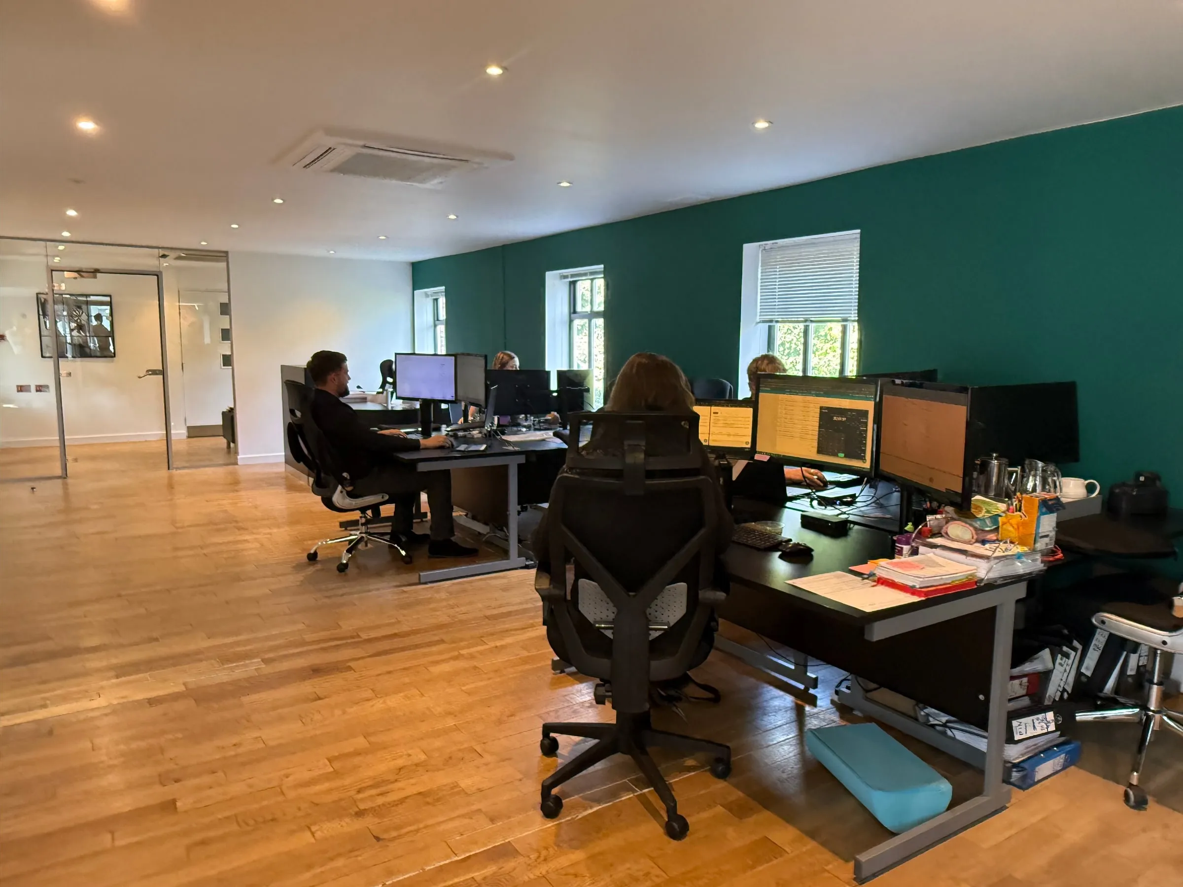 Desks along the green wall of the eba office, with daylight from the windows behind them