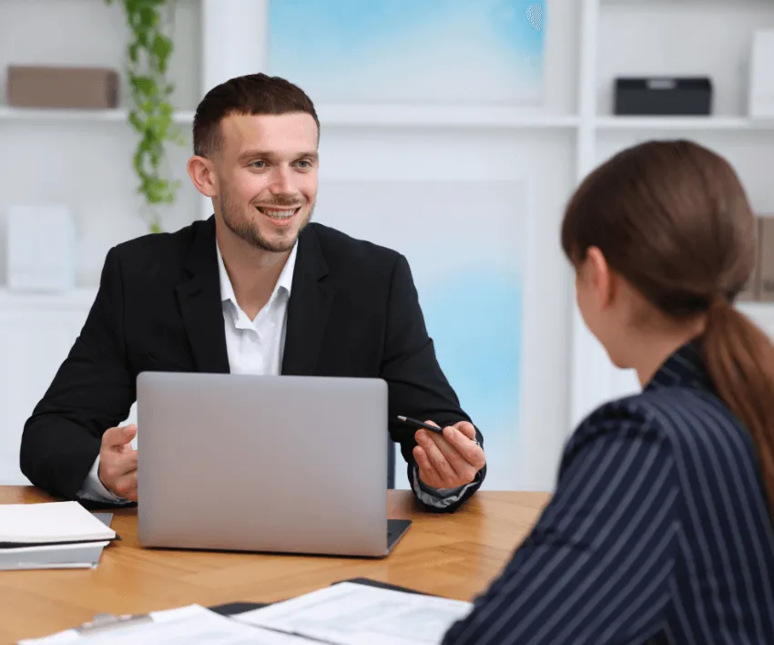 A manager and an employee talking across a desk with a laptop open between them.