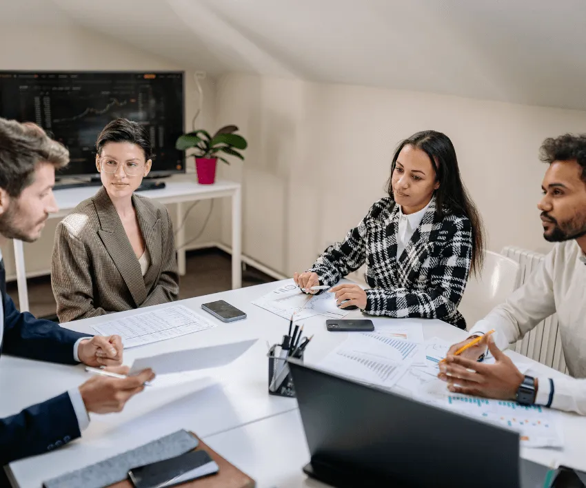 Five colleagues in a meeting around a white table with a laptop and papers in front of them.