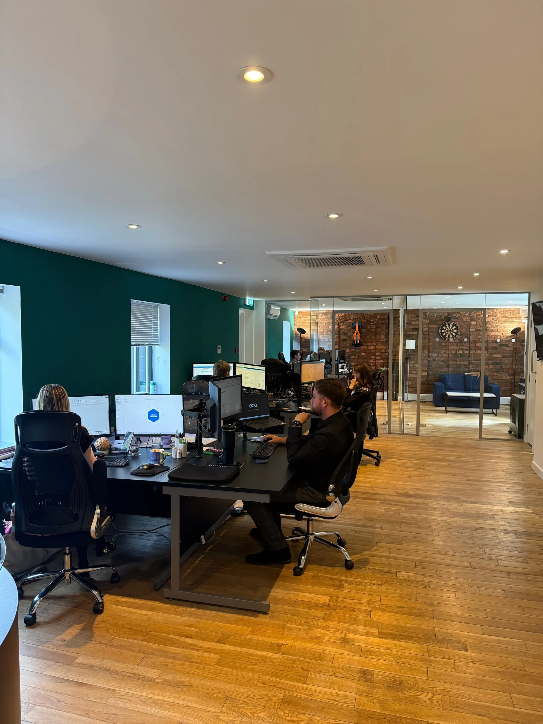 Two members of the eba team working at desks in the open plan office, with a breakout room visible beyond a glass partition
