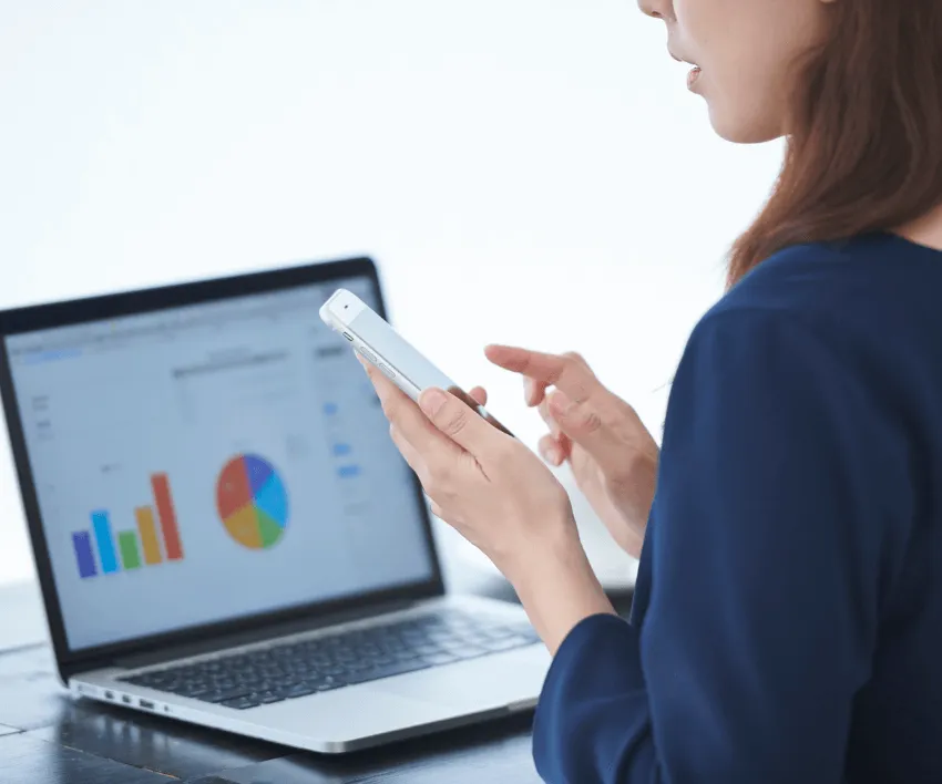 A woman holding a phone in front of a laptop showing bar and pie charts.
