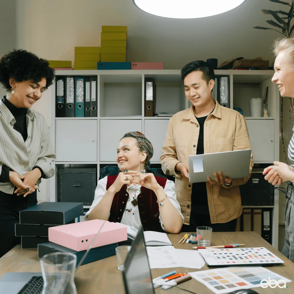 Four colleagues talking around a desk in a bright office, one of them holding a laptop.